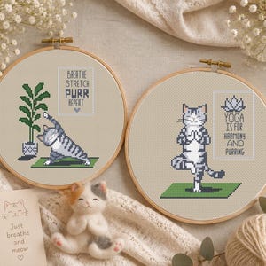 May include: Two circular wooden embroidery hoops containing cross-stitch designs of grey tabby cats practising yoga on green mats. One hoop features a cat stretching near a potted plant with the text BREATHE STRETCH PURR REPEAT. The other shows a cat in a tree pose with the text YOGA IS FOR HARMONY AND PURRING.