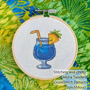 Pu&ograve; includere: Un telaio da ricamo a punto croce con un cocktail blu con cannuccia e fetta di ananas. Il disegno &egrave; ambientato su uno sfondo bianco e incorniciato da un cerchio di legno. Testo sull'immagine: Stitching and photo: Alisha Sanders, CrossStitch Pattern: StitchMood.