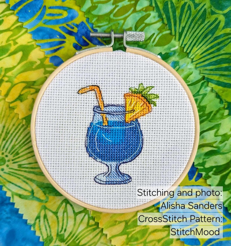Pu&ograve; includere: Un telaio da ricamo a punto croce con un cocktail blu con cannuccia e fetta di ananas. Il disegno &egrave; ambientato su uno sfondo bianco e incorniciato da un cerchio di legno. Testo sull'immagine: Stitching and photo: Alisha Sanders, CrossStitch Pattern: StitchMood.