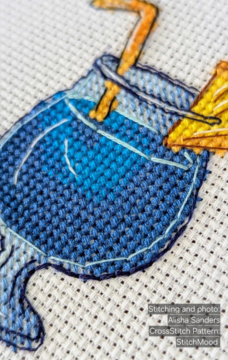Pu&ograve; includere: Primo piano di un disegno a punto croce con un cocktail blu e una guarnizione gialla. La cucitura &egrave; dettagliata, con diverse tonalit&agrave; di filo blu e giallo. Il testo "StitchMood" &egrave; visibile.