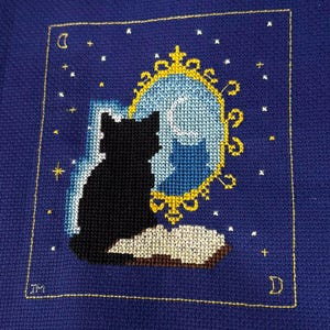 May include: Cross-stitch art featuring a black cat silhouette gazing into a mirror with a moon and stars. The mirror has a gold frame, and the background is a deep blue. The cat sits on a book, and the design is framed with a gold border.