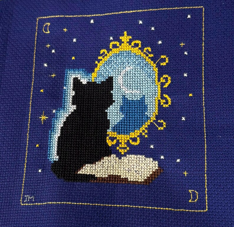 May include: Cross-stitch art featuring a black cat silhouette gazing into a mirror with a moon and stars. The mirror has a gold frame, and the background is a deep blue. The cat sits on a book, and the design is framed with a gold border.