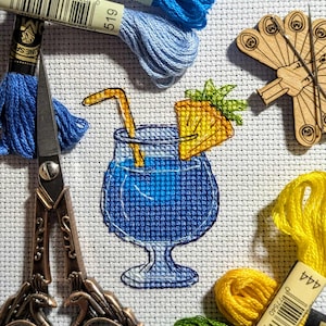 Pu&ograve; includere: Ricamo a punto croce raffigurante un cocktail blu con una fetta di ananas e una cannuccia. L'immagine include rocchetti di filo da ricamo blu, giallo e verde, un paio di forbici e un portaghiaccio in legno. Il cocktail &egrave; l'elemento centrale.
