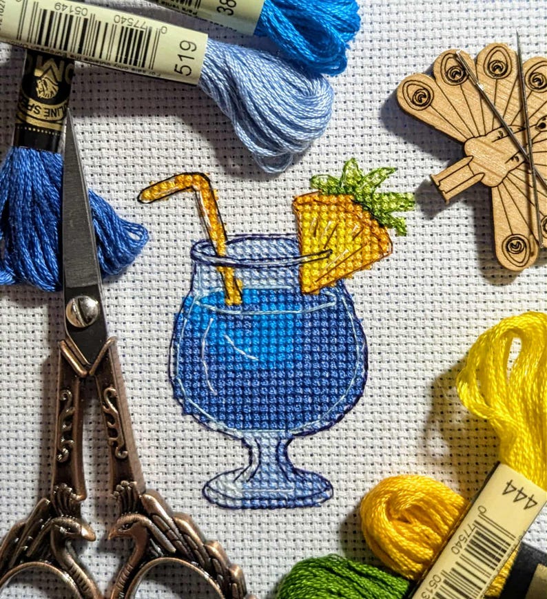 Pu&ograve; includere: Ricamo a punto croce raffigurante un cocktail blu con una fetta di ananas e una cannuccia. L'immagine include rocchetti di filo da ricamo blu, giallo e verde, un paio di forbici e un portaghiaccio in legno. Il cocktail &egrave; l'elemento centrale.