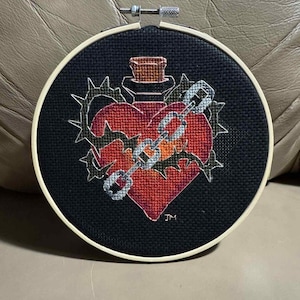 May include: A circular embroidery hoop containing black fabric with a cross-stitched design of a red heart, a glass bottle, a metal chain, and a crown of thorns. The initials JM are stitched in the lower right corner of the design.