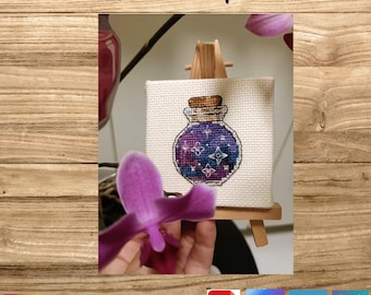 Peut inclure: Motif de point de croix d'une bouteille de potion avec un motif de nuit étoilée, exposé sur un petit chevalet en bois. La bouteille est violette et bleue avec des étoiles blanches. Le texte "Hand-drafted pattern" est en haut.