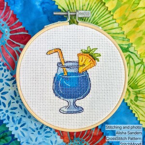 Pu&ograve; includere: Un ricamo a punto croce in un telaio di legno raffigura un cocktail blu con una cannuccia e una fetta di ananas. Il testo "StitchMood" &egrave; visibile. Lo sfondo include campioni di tessuto colorati.