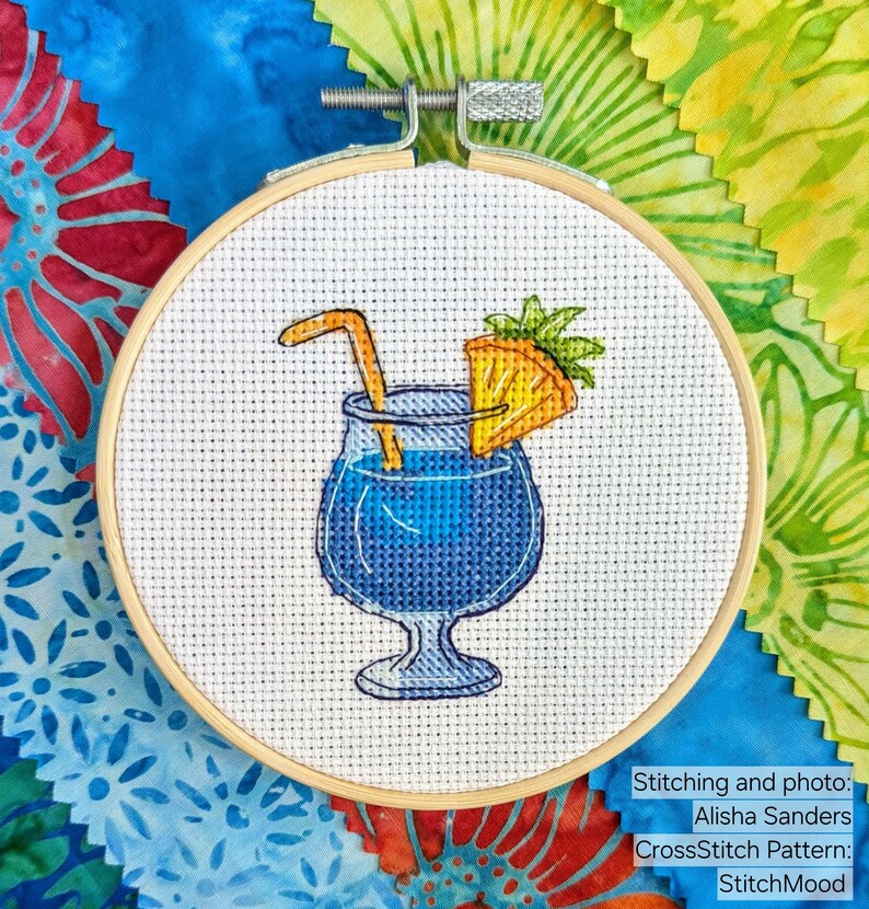 Pu&ograve; includere: Un ricamo a punto croce in un telaio di legno raffigura un cocktail blu con una cannuccia e una fetta di ananas. Il testo "StitchMood" &egrave; visibile. Lo sfondo include campioni di tessuto colorati.