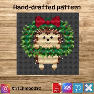 Puede incluir: Patrón de punto de cruz con un erizo en una corona navideña con un lazo rojo. El diseño está sobre un fondo gris oscuro con el texto "Hand-drafted pattern" en la parte superior. La imagen también incluye el nombre de usuario de Instagram "Stitchmood92".