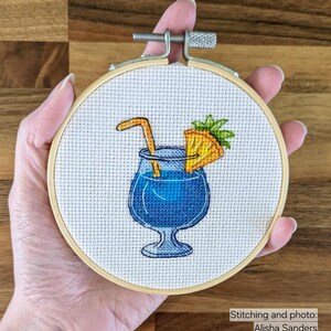 Pu&ograve; includere: Ricamo a punto croce in un telaio di legno. Il disegno presenta un cocktail blu con una cannuccia e una fetta d'arancia. Il testo "StitchMood" &egrave; visibile nell'immagine.