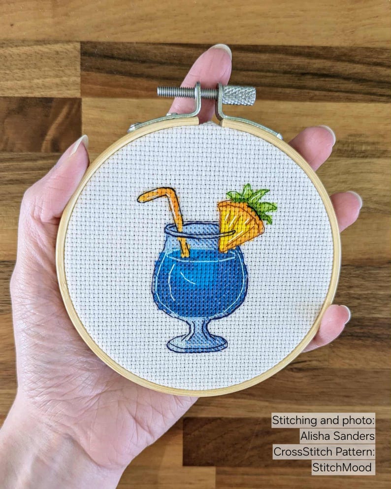 Pu&ograve; includere: Ricamo a punto croce in un telaio di legno. Il disegno presenta un cocktail blu con una cannuccia e una fetta d'arancia. Il testo "StitchMood" &egrave; visibile nell'immagine.