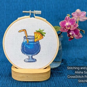 Pu&ograve; includere: Un ricamo a punto croce di un cocktail blu con una guarnizione di ananas e una cannuccia, in un telaio di legno. Il ricamo &egrave; esposto su un supporto di legno, con uno sfondo blu e un vaso di orchidee rosa.
