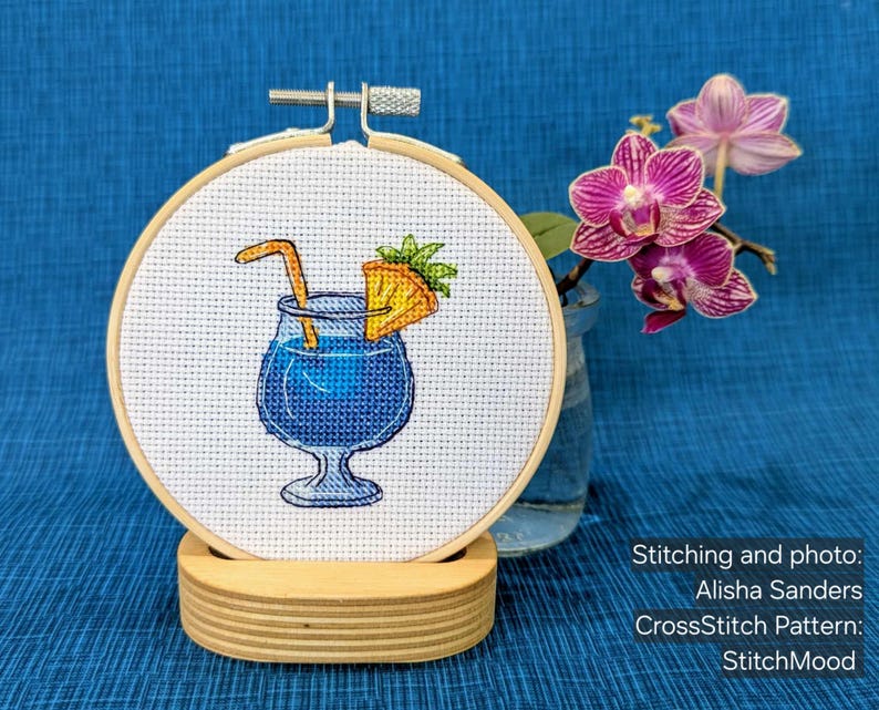 Pu&ograve; includere: Un ricamo a punto croce di un cocktail blu con una guarnizione di ananas e una cannuccia, in un telaio di legno. Il ricamo &egrave; esposto su un supporto di legno, con uno sfondo blu e un vaso di orchidee rosa.