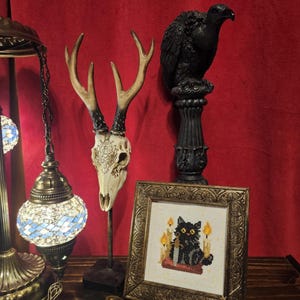 May include: A decorative display featuring a deer skull with antlers, a black bird statue on a pedestal, and a framed cross-stitch of a black cat with candles. The background is a deep red curtain, and the items are on a wooden surface.