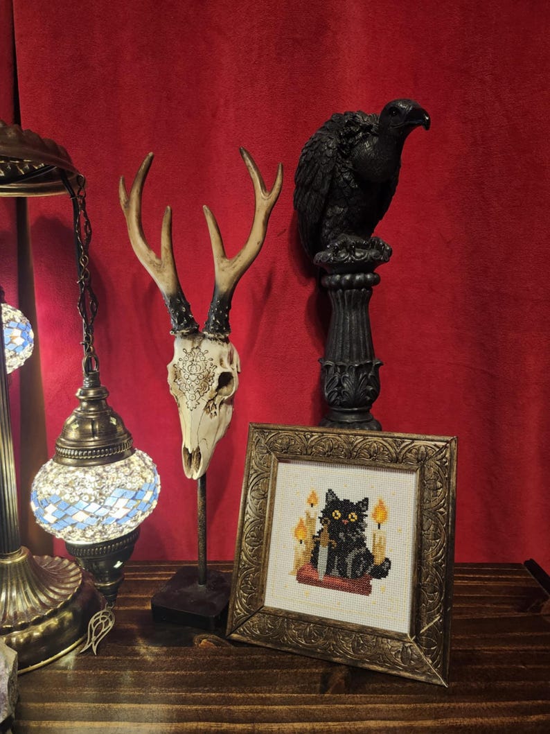 May include: A decorative display featuring a deer skull with antlers, a black bird statue on a pedestal, and a framed cross-stitch of a black cat with candles. The background is a deep red curtain, and the items are on a wooden surface.