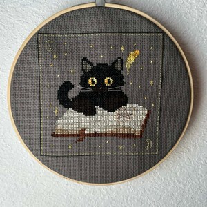 May include: Cross-stitch embroidery of a black cat with yellow eyes sitting on an open book. The cat is surrounded by gold stars and a gold feather. The embroidery is set in a gray fabric within a wooden hoop.
