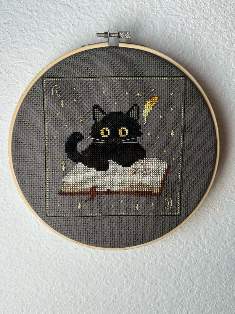 May include: Cross-stitch embroidery of a black cat with yellow eyes sitting on an open book. The cat is surrounded by gold stars and a gold feather. The embroidery is set in a gray fabric within a wooden hoop.