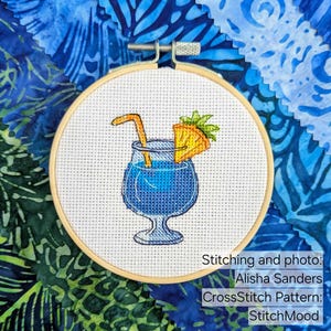 Pu&ograve; includere: Ricamo a punto croce di un cocktail blu con guarnizione di ananas e cannuccia, in un cerchio di legno. Lo sfondo presenta tessuto blu con vari motivi. Testo: Stitching and photo: Alisha Sanders, CrossStitch Pattern: StitchMood.