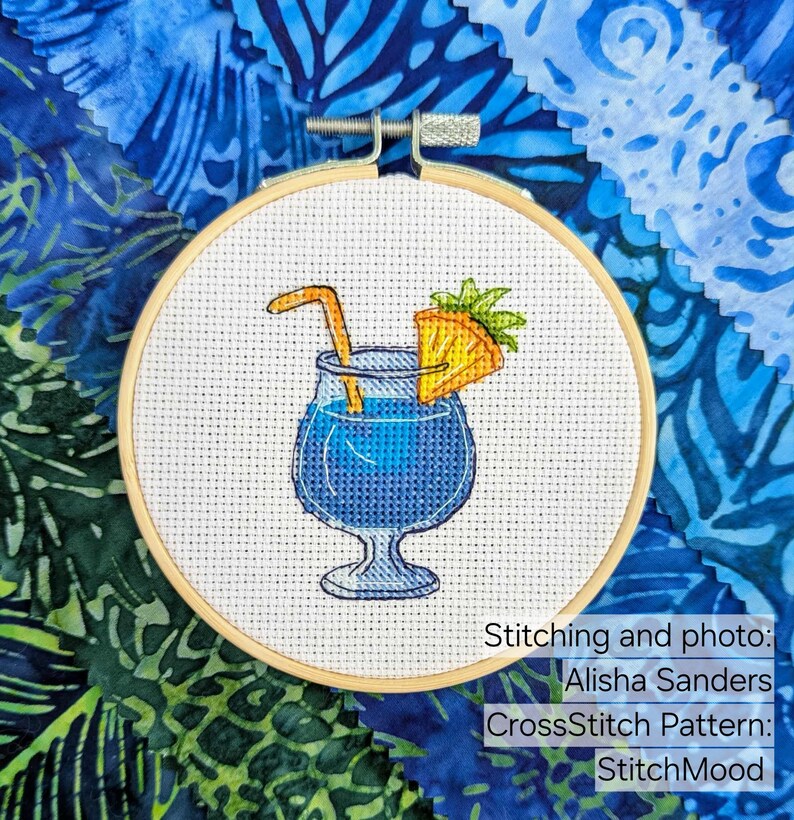 Pu&ograve; includere: Ricamo a punto croce di un cocktail blu con guarnizione di ananas e cannuccia, in un cerchio di legno. Lo sfondo presenta tessuto blu con vari motivi. Testo: Stitching and photo: Alisha Sanders, CrossStitch Pattern: StitchMood.