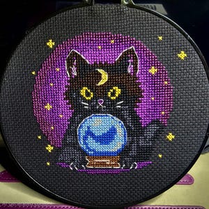 May include: Cross-stitch embroidery in a black hoop. The design features a black cat with yellow eyes and a crescent moon on its forehead, gazing into a blue crystal ball. A purple moon and yellow stars complete the mystical scene.