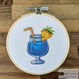 Pu&ograve; includere: Ricamo a punto croce di un cocktail blu con guarnizione di ananas e cannuccia, in un telaio da ricamo in legno. Il testo "StitchMood" &egrave; visibile.