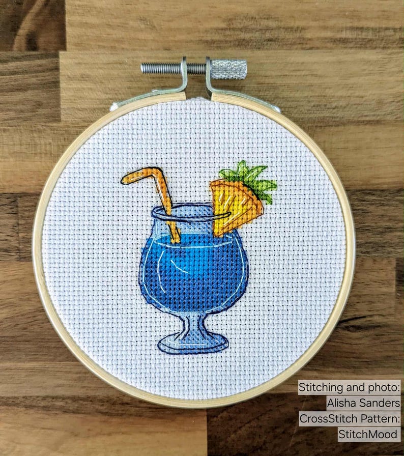 Pu&ograve; includere: Ricamo a punto croce di un cocktail blu con guarnizione di ananas e cannuccia, in un telaio da ricamo in legno. Il testo "StitchMood" &egrave; visibile.
