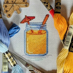 Op de afbeelding: Een kruissteekontwerp van een cocktail in een pot met een rode paraplu en een rietje. De afbeelding is omgeven door borduurgaren in blauwe, oranje en gele tinten, samen met een schaar en een houten pauw-naaldhouder.