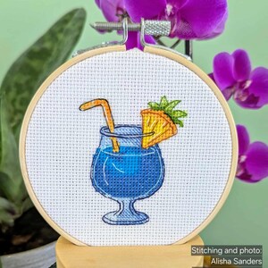Pu&ograve; includere: Ricamo a punto croce in un telaio di legno raffigurante un cocktail blu con una fetta di ananas e una cannuccia. Il ricamo &egrave; ambientato su uno sfondo di orchidee viola e foglie verdi. Il testo "Stitching and photo: Alisha Sanders CrossStitch Pattern: StitchMood" &egrave; visibile.