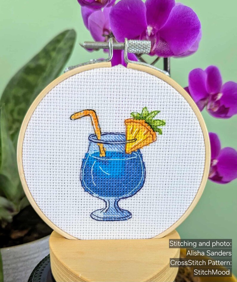 Pu&ograve; includere: Ricamo a punto croce in un telaio di legno raffigurante un cocktail blu con una fetta di ananas e una cannuccia. Il ricamo &egrave; ambientato su uno sfondo di orchidee viola e foglie verdi. Il testo "Stitching and photo: Alisha Sanders CrossStitch Pattern: StitchMood" &egrave; visibile.