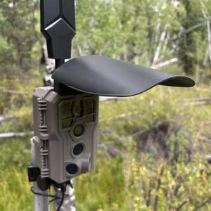 May include: A trail camera with a light gray rectangular housing, featuring a central lens, infrared sensors, and a black plastic sunshade mounted above. The device is attached to a vertical metal pole with a black antenna extending upward.