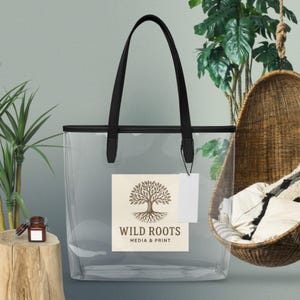 May include: Clear tote bag with black handles and trim. The bag features a rectangular label with the text "WILD ROOTS MEDIA & PRINT" and a tree logo. A blank tag hangs from the handle. The bag is transparent.