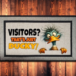 May include: Rectangular doormat with a gray border and a cartoon duck illustration. The text reads "VISITORS? THAT'S JUST DUCKY!" in black and orange. The background is a wood grain pattern.