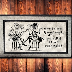 May include: Framed artwork featuring a black and white illustration of two women seated on stools, holding cups. The text reads, "remember dear if we get caught... you're blind & I don't speak english!" The artwork is framed with a gray and black border.