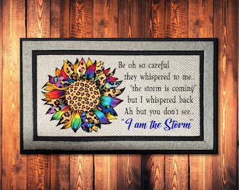May include: A framed canvas print featuring a vibrant sunflower with rainbow-colored petals and a leopard print center. The text reads "I am the Storm" in blue script, with additional text in black. The frame is black with a gray border, set against a wood background.