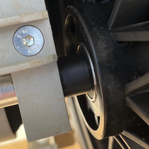 May include: Close-up of a mechanical component, featuring a black cylindrical part with ribbed texture, connected to a black wheel. A silver bolt secures the component to a light gray metal piece.