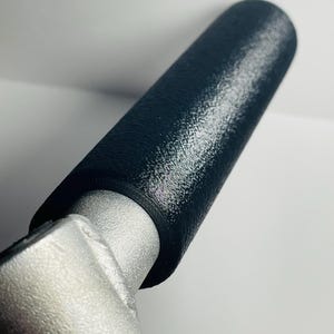 May include: Close-up of a black textured handle grip attached to a silver metal component. The handle is cylindrical and appears to be made of a rubberized material. The metal component has a matte finish.