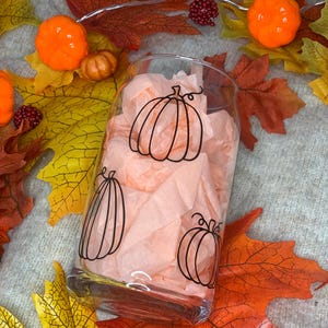 May include: A clear glass with a black outline of pumpkins on it. The glass is filled with peach-colored tissue paper. The glass is sitting on a surface with fall leaves.