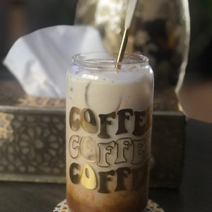 May include: A glass of iced coffee with a gold straw in a clear glass with the word "COFFEE" repeated three times in gold letters on the side. The glass is sitting on a coaster with a geometric pattern.