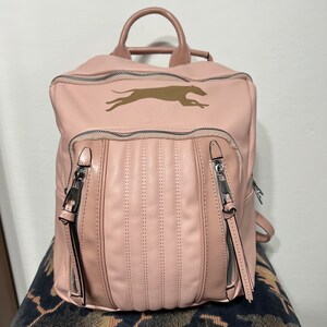 以下が含まれることがあります： ピンク色の革製バックパック。上部にハンドルがあり、前面に2つのジッパー付きポケットが付いています。バックパックには、金色の犬のロゴと、前面に垂直ステッチのディテールがあります。ジッパーには長い黒いプルが付いています。