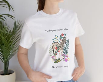 May include: A white short-sleeved t-shirt featuring a graphic of a zebra surrounded by colorful flowers and green leaves. The text above the zebra reads Finding out you're a zebra and the text below the zebra reads Not a "weird" horse.