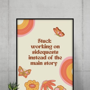 May include: A framed art print with a retro design. The print features the text "Stuck working on sidequests instead of the main story" in brown font. It includes illustrations of flowers, butterflies, and a rainbow-colored circle.
