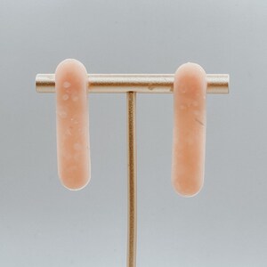 Op de afbeelding: Een paar lange, rechthoekige oorbellen in perzikkleur met een gespikkelde textuur. De oorbellen worden gepresenteerd op een gouden standaard.