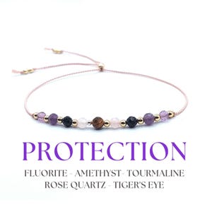 May include: A pink string bracelet with beads of fluorite, amethyst, tourmaline, rose quartz, and tiger's eye. The bracelet is tied with a knot and has gold beads. The bracelet is labeled "PROTECTION" and lists the stones used: "FLUORITE - AMETHYST - TOURMALINE ROSE QUARTZ - TIGER'S EYE".