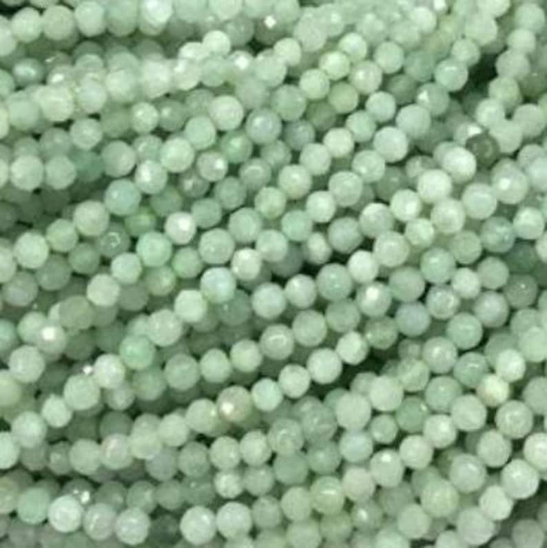 May include: A collection of small, round, faceted beads in a pale mint green color. Each bead features a multi-faceted surface that reflects light, creating a subtle shimmer across the dense arrangement of these uniform, spherical craft components.