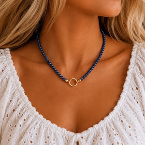 May include: A necklace featuring a strand of small, round, deep blue beads. The beads are connected at the centre by a polished gold-tone circular ring clasp. The necklace rests against the neck, showcasing a uniform bead size and a smooth finish.