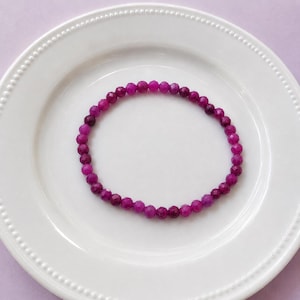 Puede incluir: Una pulsera circular compuesta por pequeñas cuentas facetadas de color magenta intenso. Las cuentas son uniformes en tamaño y forma, creando un bucle continuo. La superficie de cada cuenta refleja la luz debido al corte multifacetado.