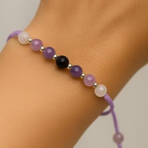 May include: A beaded bracelet featuring a string of purple, white, and black gemstones. The bracelet is strung on a purple cord and has silver-colored metal beads between each gemstone. The bracelet is on a wrist.