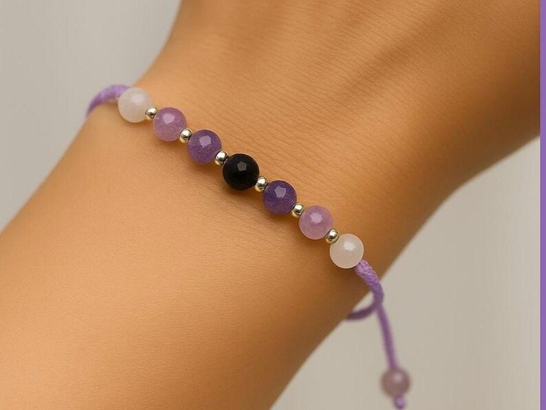 May include: A beaded bracelet featuring a string of purple, white, and black gemstones. The bracelet is strung on a purple cord and has silver-colored metal beads between each gemstone. The bracelet is on a wrist.