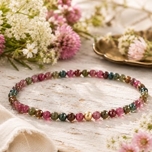 May include: A beaded necklace featuring small, round, multi-colored tourmaline gemstones in shades of pink, green, and brown, accented by a single smooth, polished gold-toned spherical bead positioned near the center of the circular strand.