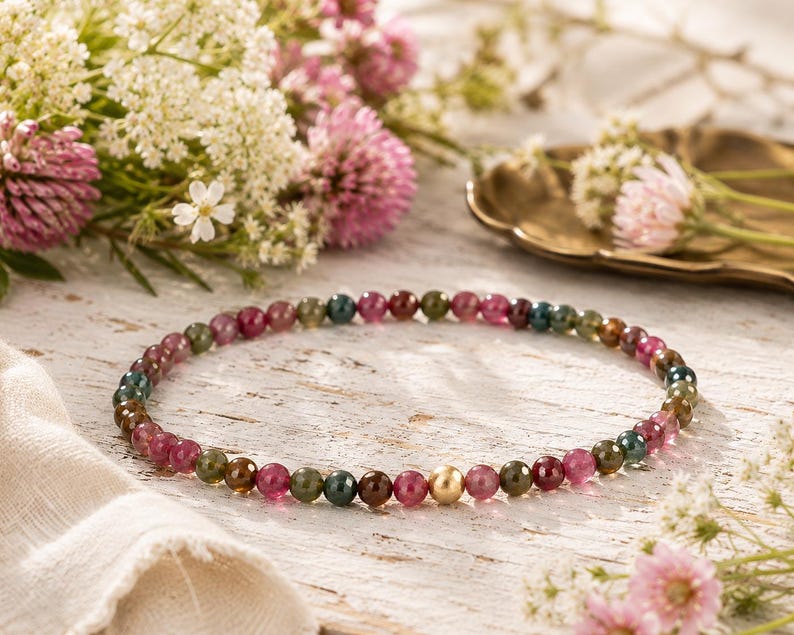May include: A beaded necklace featuring small, round, multi-colored tourmaline gemstones in shades of pink, green, and brown, accented by a single smooth, polished gold-toned spherical bead positioned near the center of the circular strand.